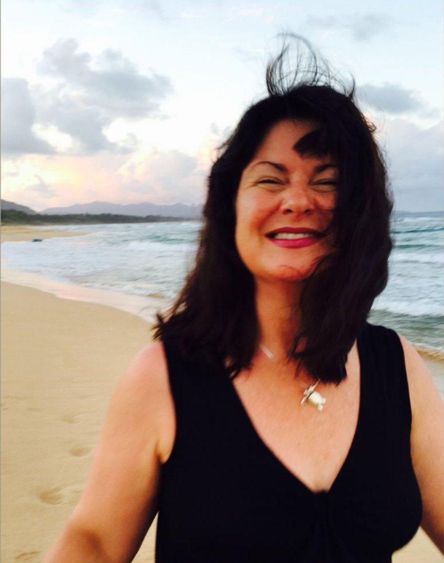 Katie Rose smiling on a sandy beach at sunset, ocean and mountains behind her